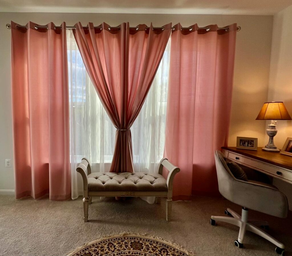 pexels-photo-36881833 Cozy interior featuring soft pink curtains, a tufted bench, and a stylish desk with lamp.